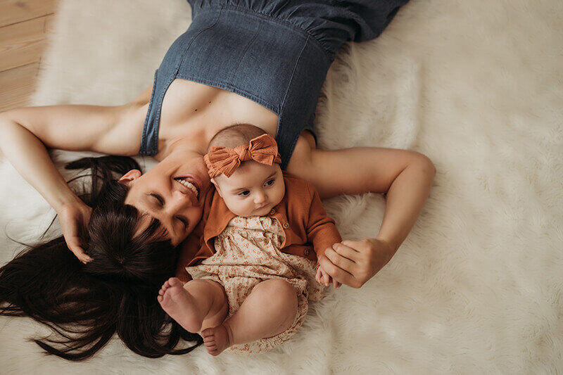 Mom in denim dress lays on her back with four month old baby girl in orange floral dress
