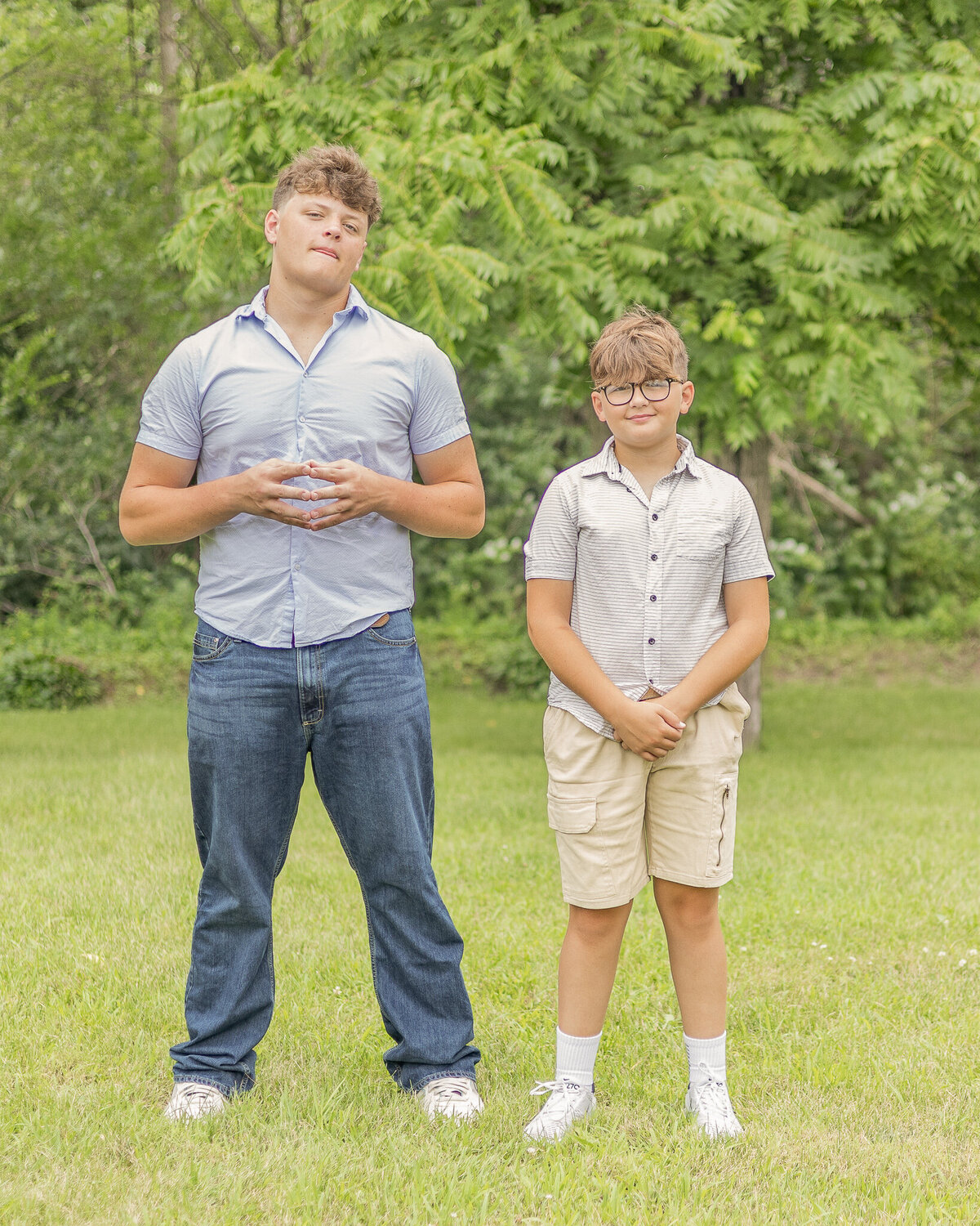 Two brothers standing next to each other on grass with a tree line behind them.  Older brother is looking at the camera and the other brother is looking at the camera with a smile.  Both have short hair.