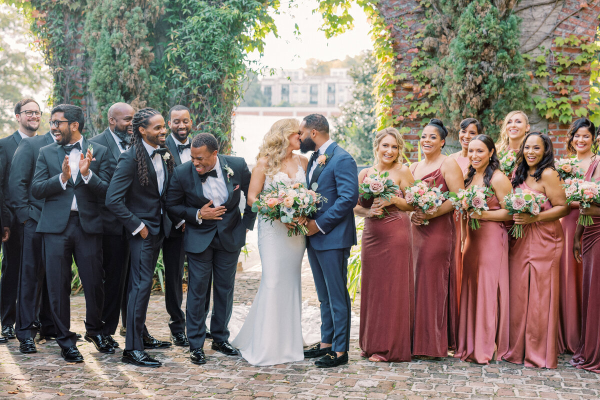 Bride and groom kiss under ivy filled archway with wedding party cheering at Summerour Studio in Georgia