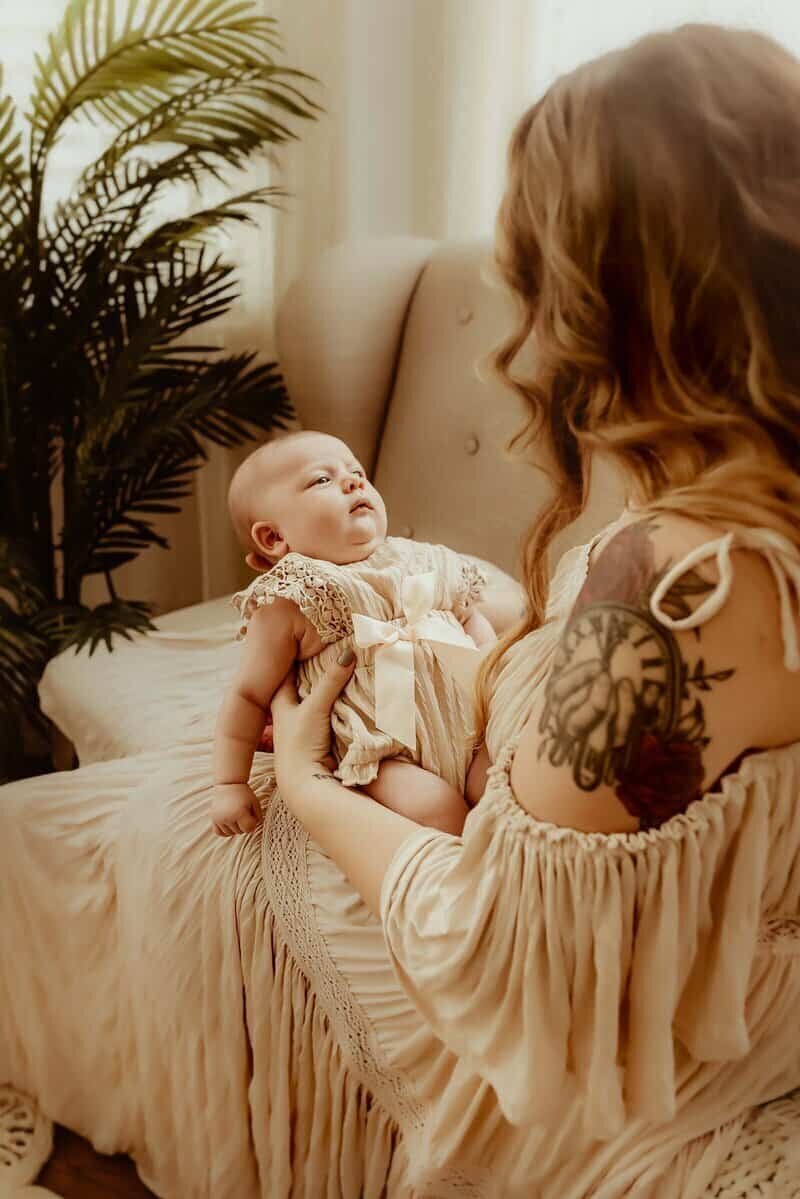 A woman with a floral tattoo on her arm holds a baby in a cream dress while sitting on a beige chair in a softly lit room with a plant in the background.