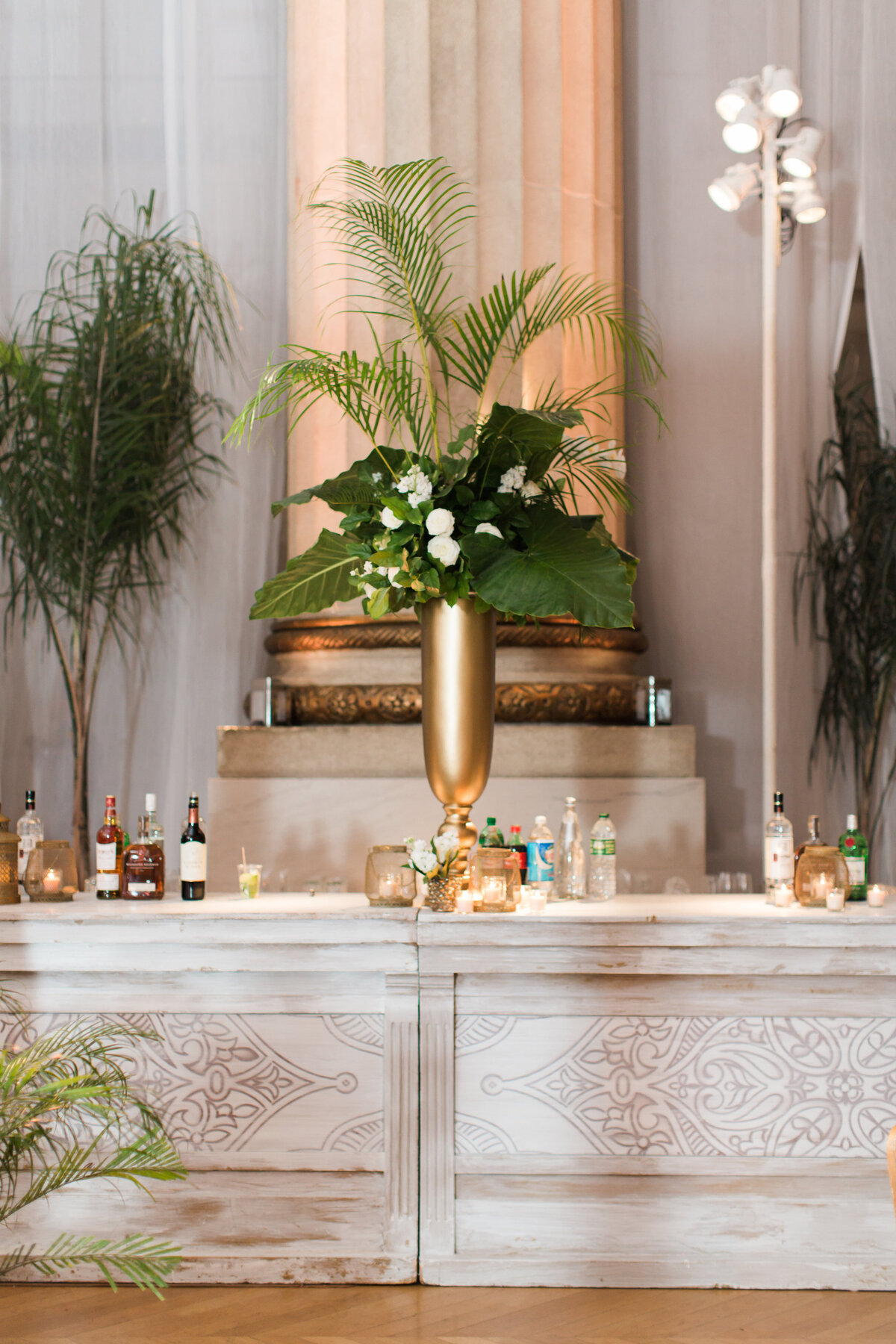 The photo was taken at the Mellon Auditorium in Washington D.C., known as one of the best luxury DC venues with European flair, captured by photographer Sarah Bradshaw. In the image, there is a decorative bar setup featuring a large golden vase filled with tropical green leaves and white flowers placed on a white counter. Neatly arranged bottles and glasses can be seen on the counter, with tall columns and additional green plants visible in the background.