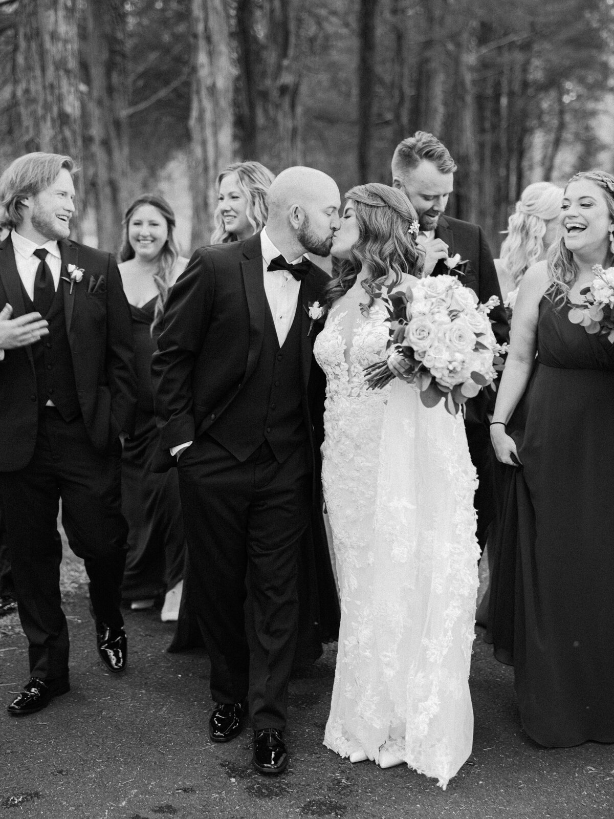 A black and white image of a bride and groom sharing a kiss as they walk with their bridal party
