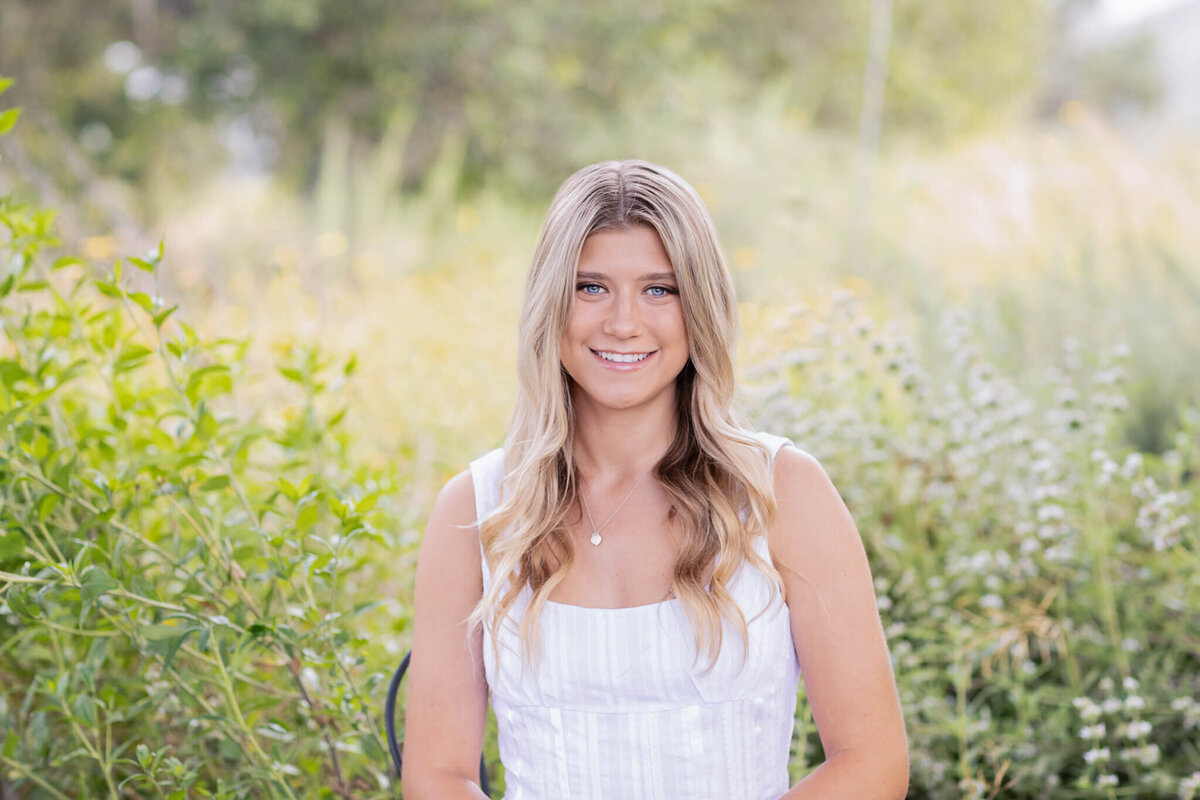 A young woman with long, wavy blonde hair is smiling while sitting outdoors. She is wearing a white sleeveless top, surrounded by green foliage and blurred plants in the background.