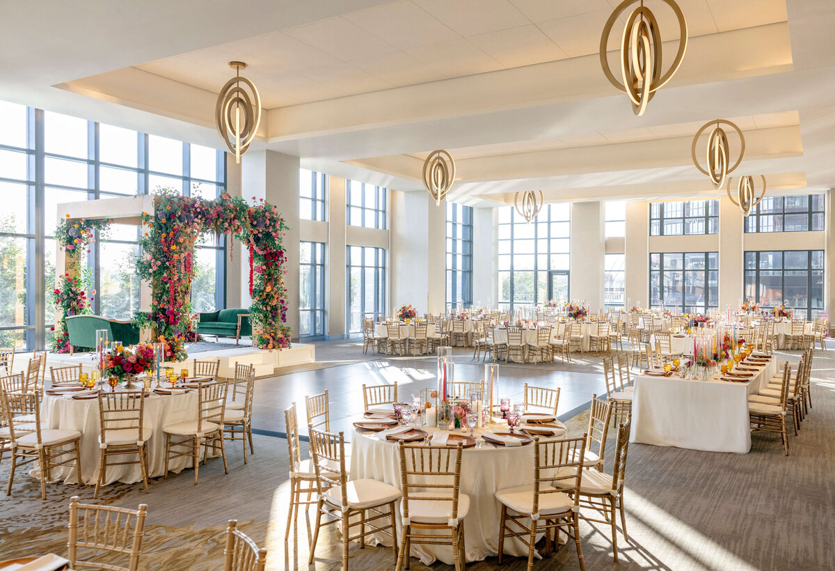 Spacious wedding reception hall with elegant round tables, white tablecloths, and gold chairs. Vibrant floral arch on a raised stage and large windows letting in natural light. Modern chandeliers hang from the ceiling.