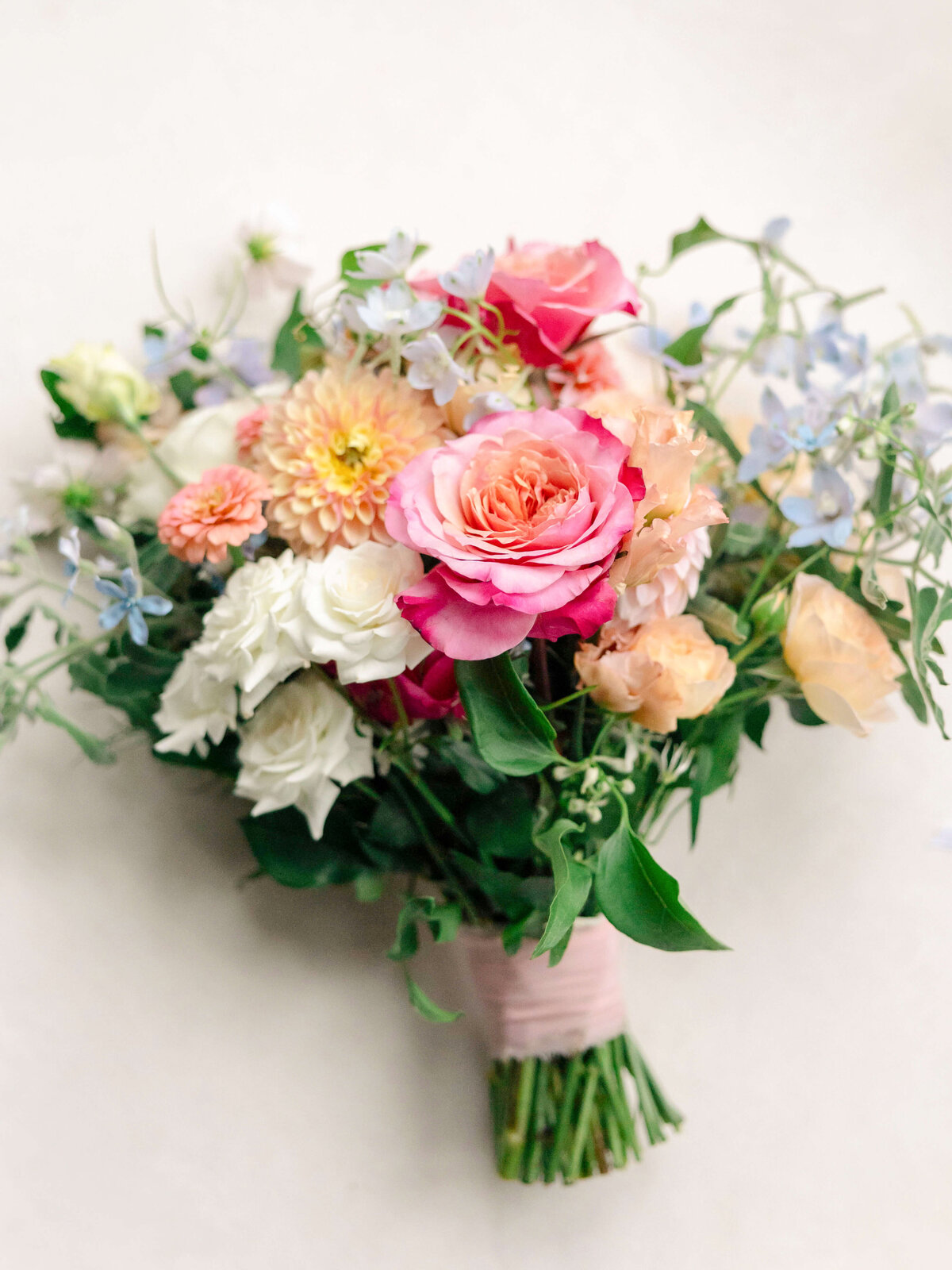 A colorful bouquet featuring pink roses, white and peach flowers, and scattered light blue blooms is arranged against a neutral background, with green leaves adding a touch of freshness.