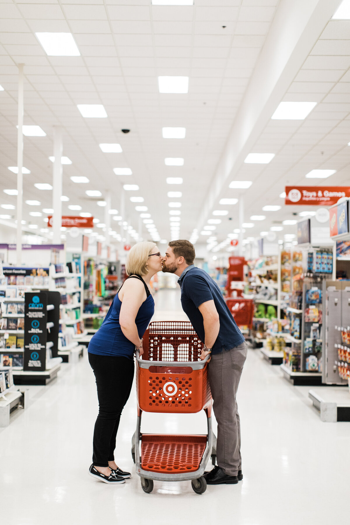 A couple sharing a kiss over a shopping cart at a Target during their engagement session in DFW, Texas. The woman on the right is wearing a dark blue tank top, black pants, and glasses. The man on the left is wearing a short sleeve collared blue shirt and grey pants. Many rows of the Target store can be seen in the background.