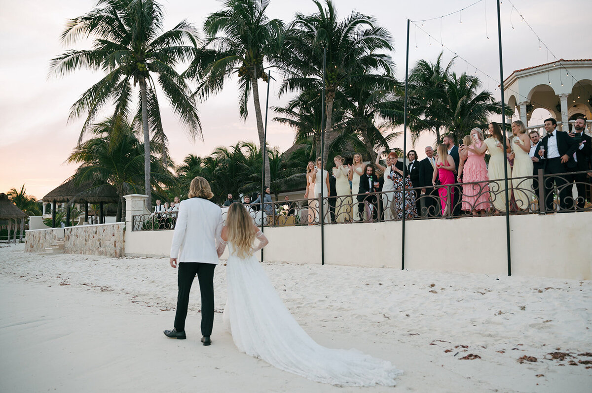 married couple standing on beach with back to camera looking at the water with a pier extending from the right