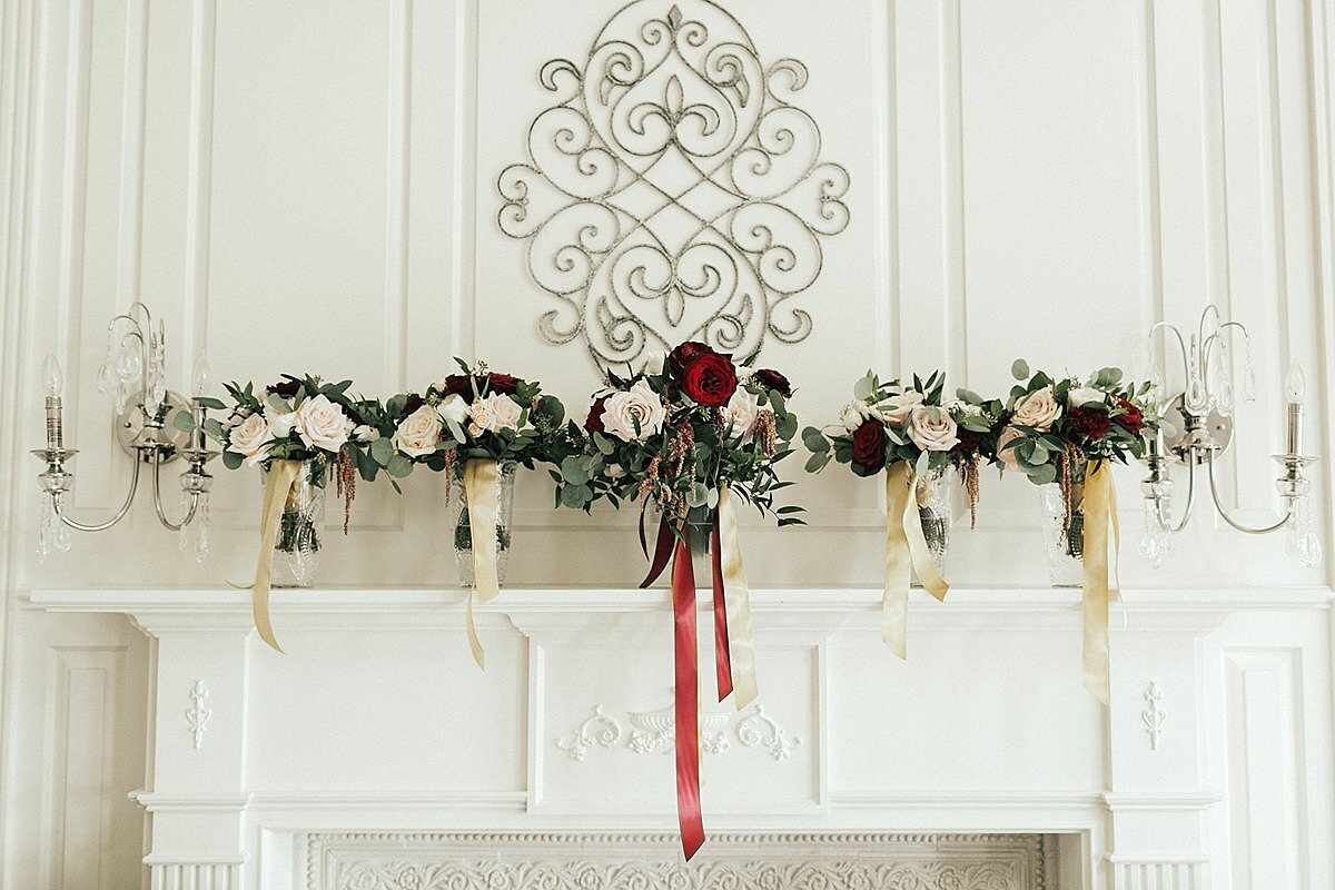 A white mantle on a white shiplap wall with a filagree wall hanging above a row of round white bridesmaid bouquets with white satin ribbons sit on either side