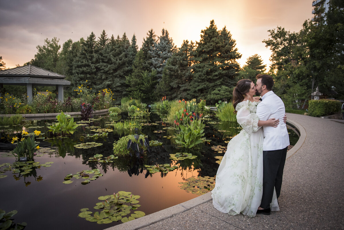 A bride and groom share a kiss in front of a small pond with lily pads.