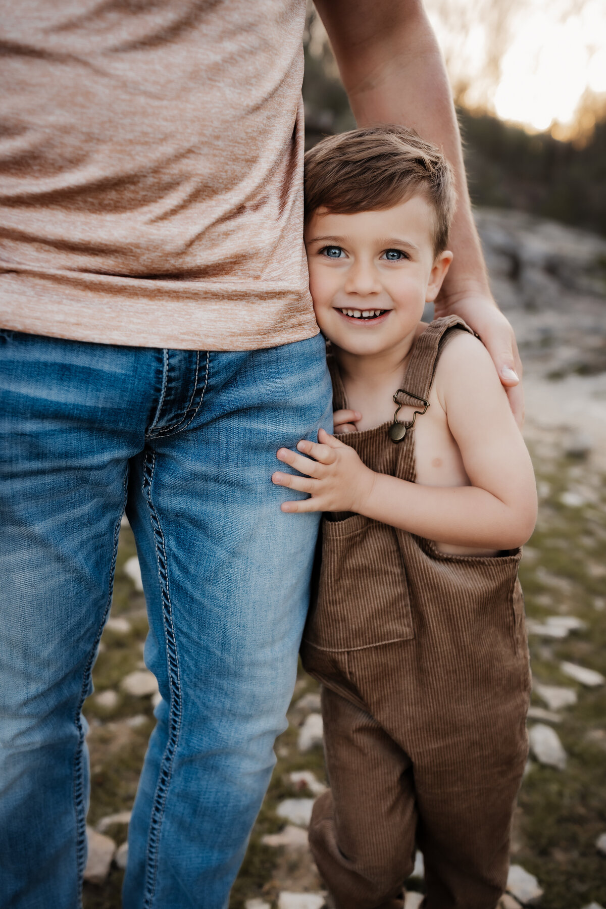 boy in borwn overalls hugging dads leg