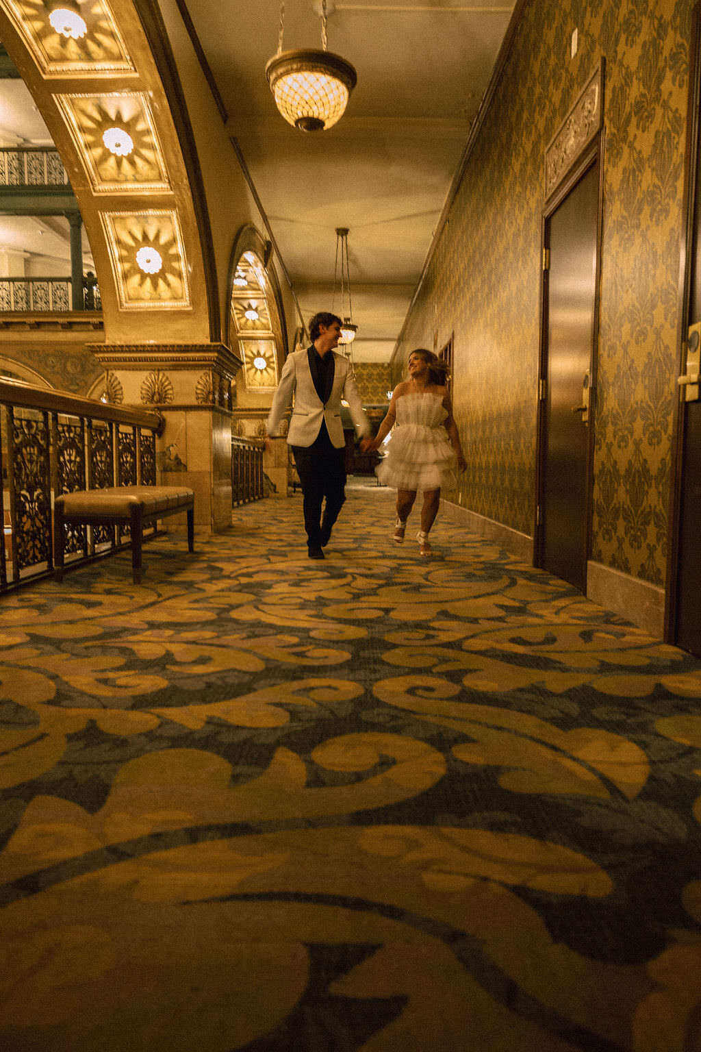 A couple holding hands and walking down a hotel hallway.