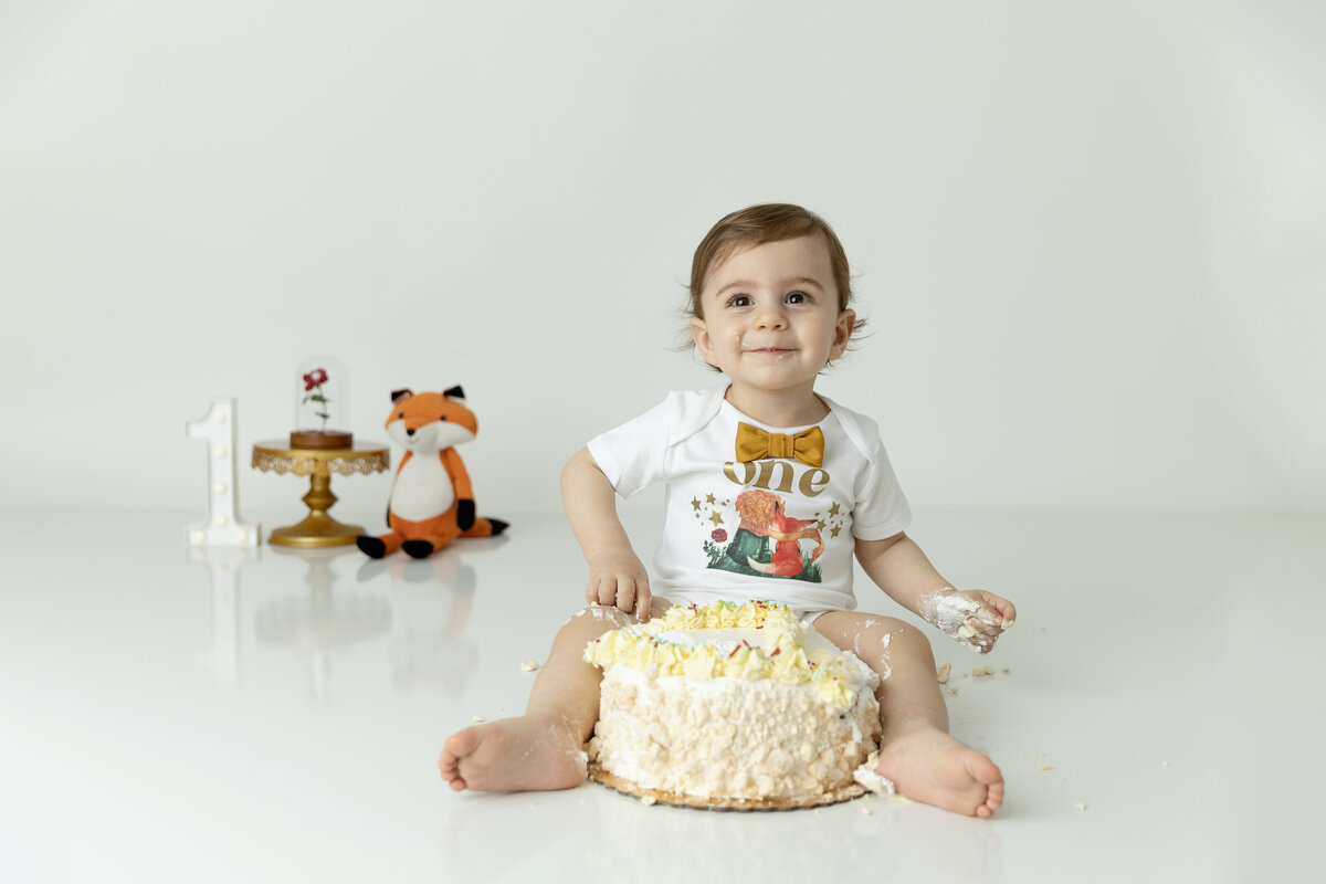 A toddler boy sits on the floor of a studio with a frosted cake between his legs for his first birthday