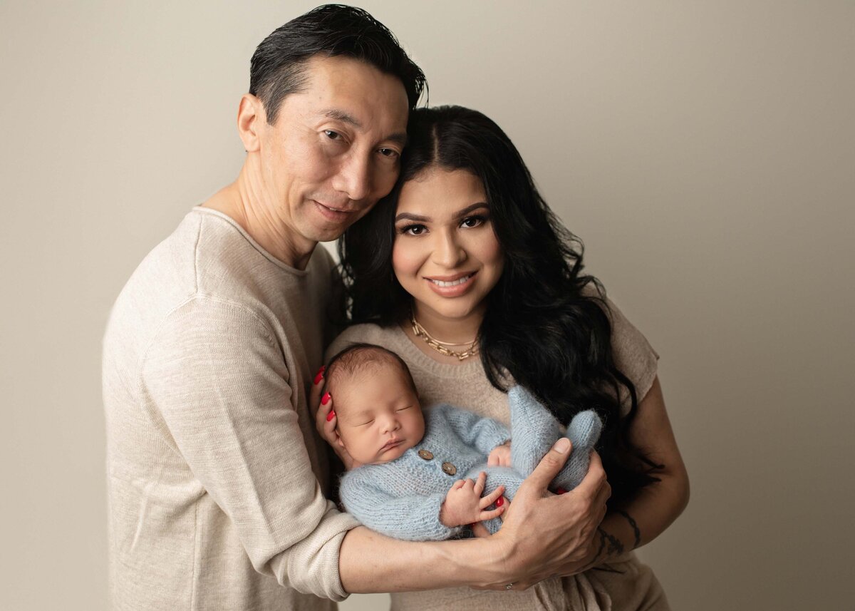 Mom and dad pose with their newborn baby boy for their Menifee newborn photoshoot. Mom is holding the baby and dad is standing slightly behind mom. Mom and dad have their heads resting together and smiling at the camera. Captured by best Menifee newborn photographer Bonny Lynn Photography