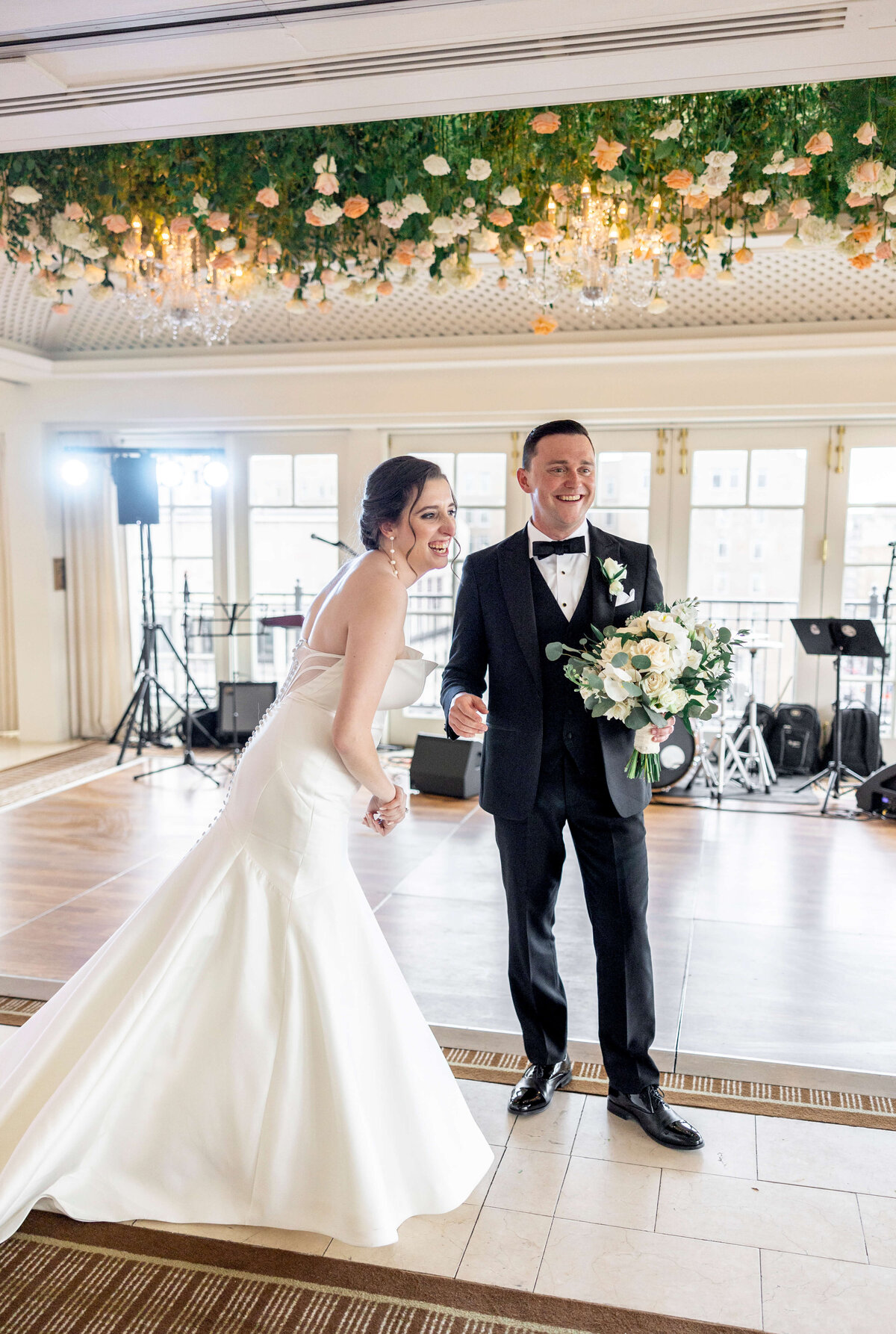 A bride and groom smile joyously, standing side by side. The bride is in a white gown and holding her dress, while the groom wears a black suit and holds a bouquet. Theyre in a decorated venue with floral fixtures and musical equipment in the background.