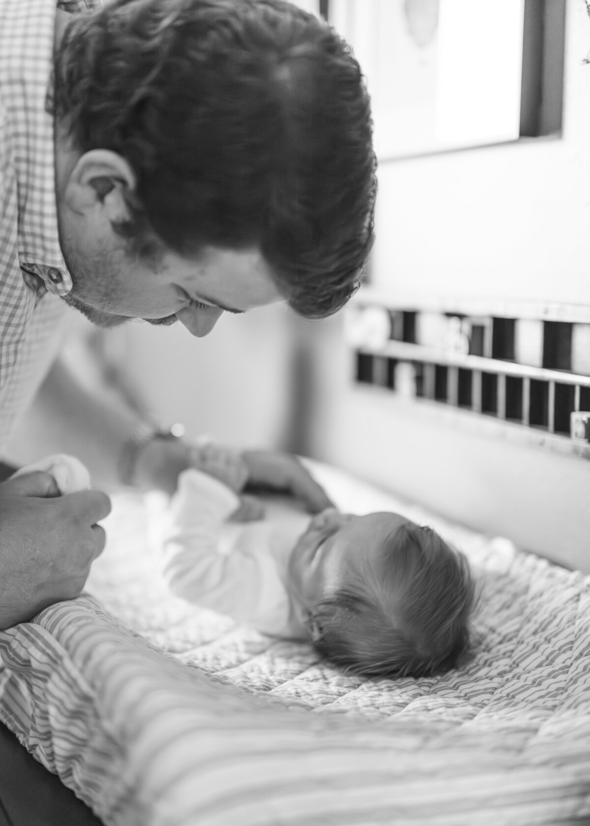 Black and white image of a father kissing his newborn as they lay in a crib, captured in an in-home Kansas City family photography session.