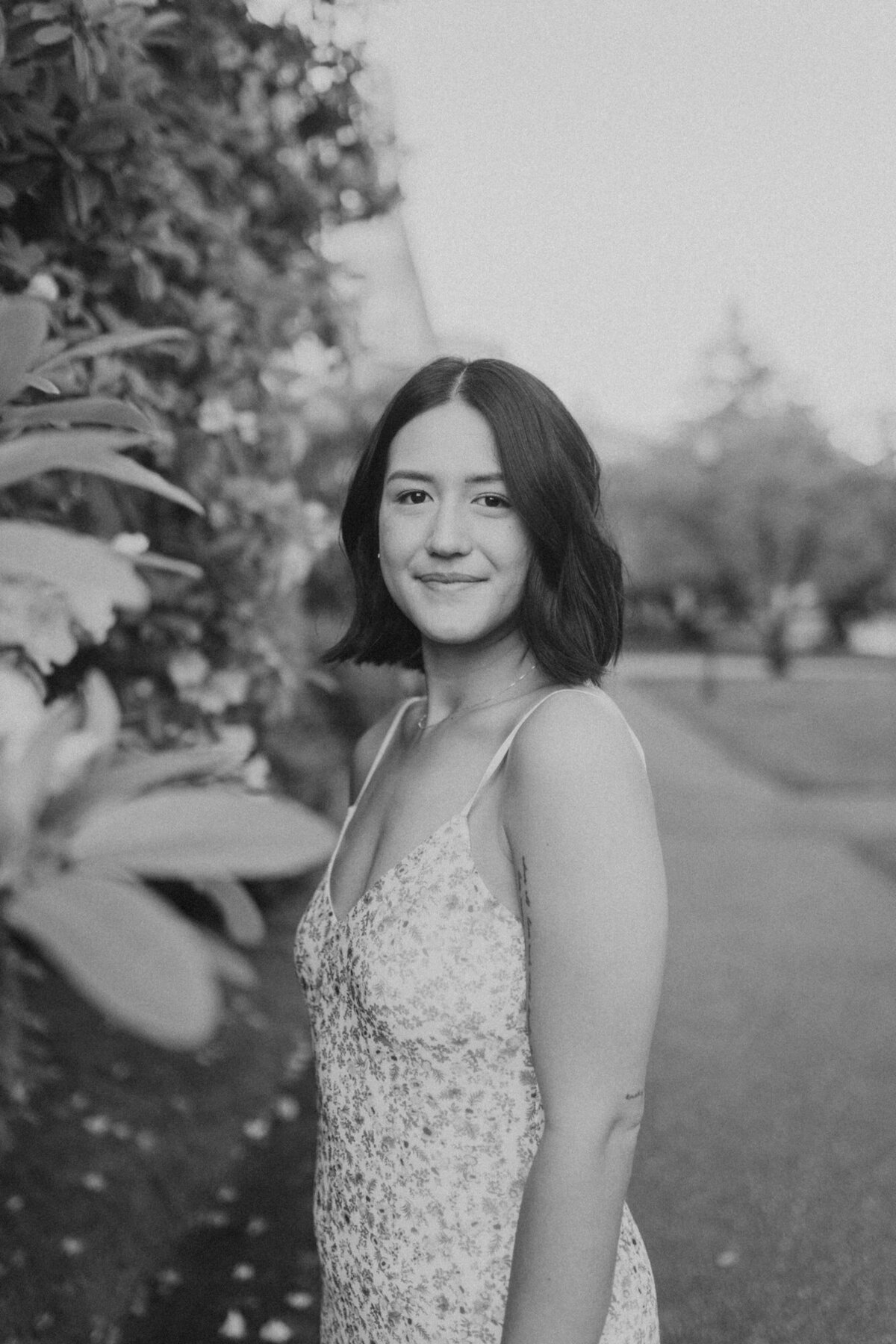 Black and white image of girl looking at camera while standing next to a bush