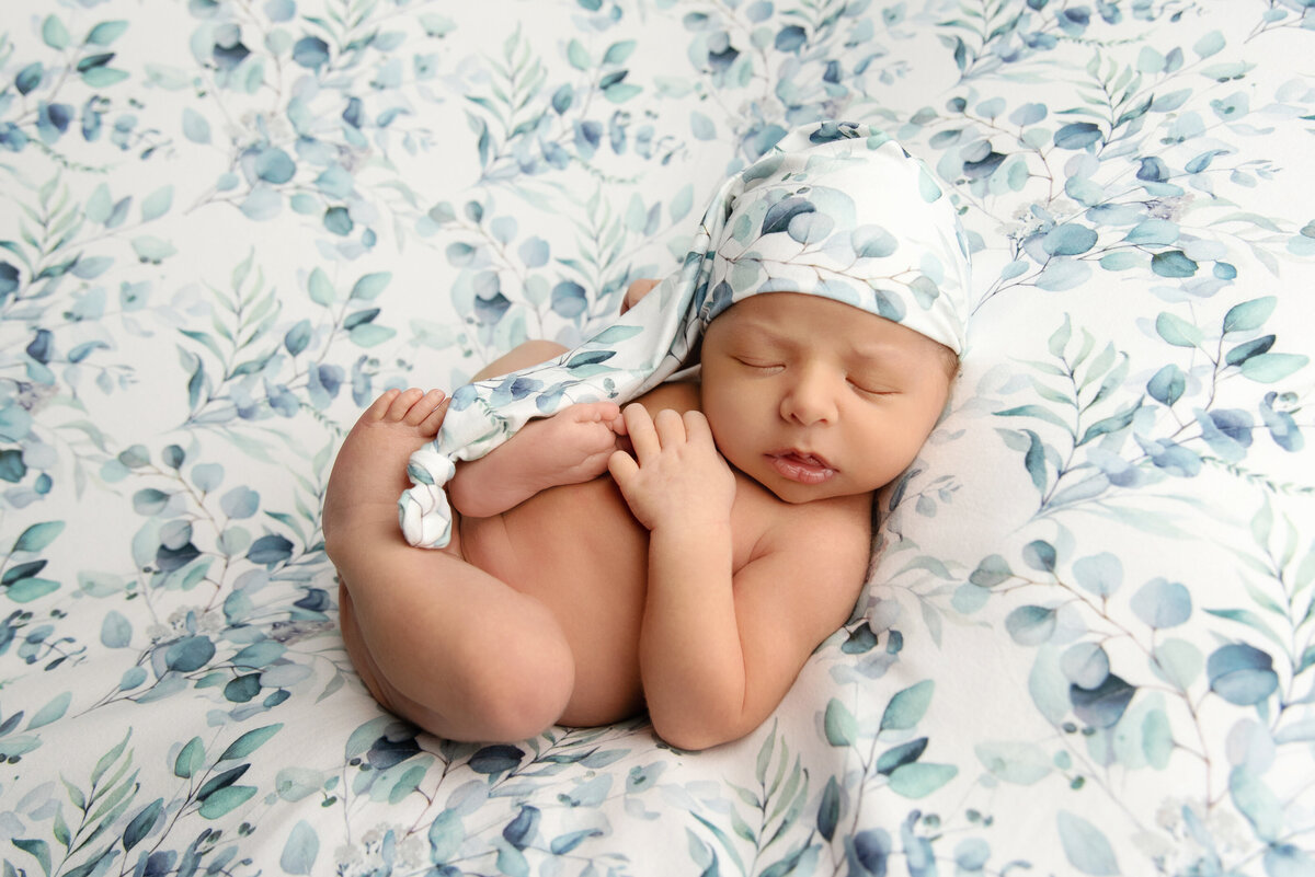 st-louis-newborn-photographer-baby-boy-wearing-sleepy-hat-laying-on-blue-green-leaf-backdrop