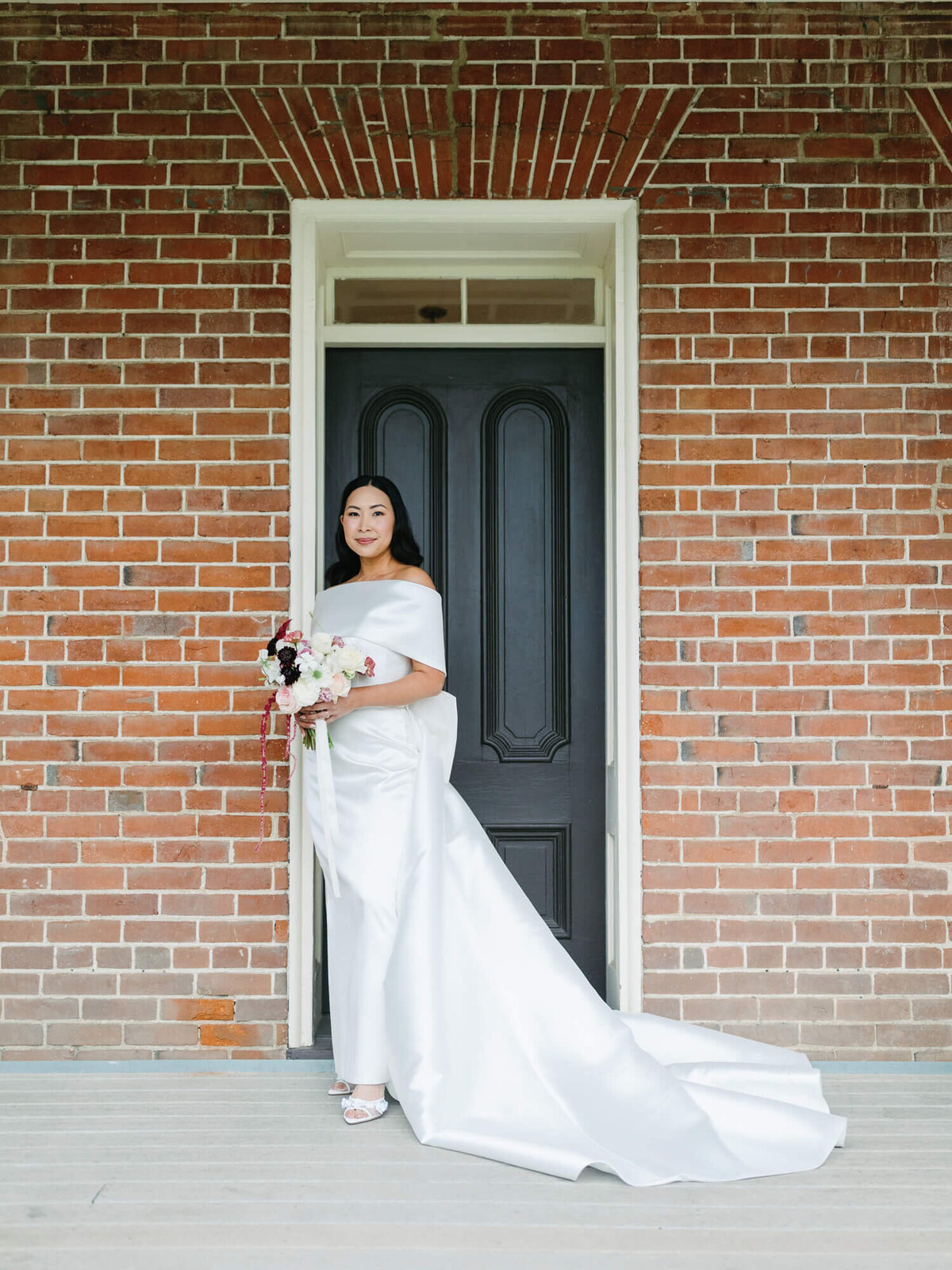 A bride in an off-shoulder white gown stands in front of a black door, holding a bouquet of flowers, framed by a brick wall.