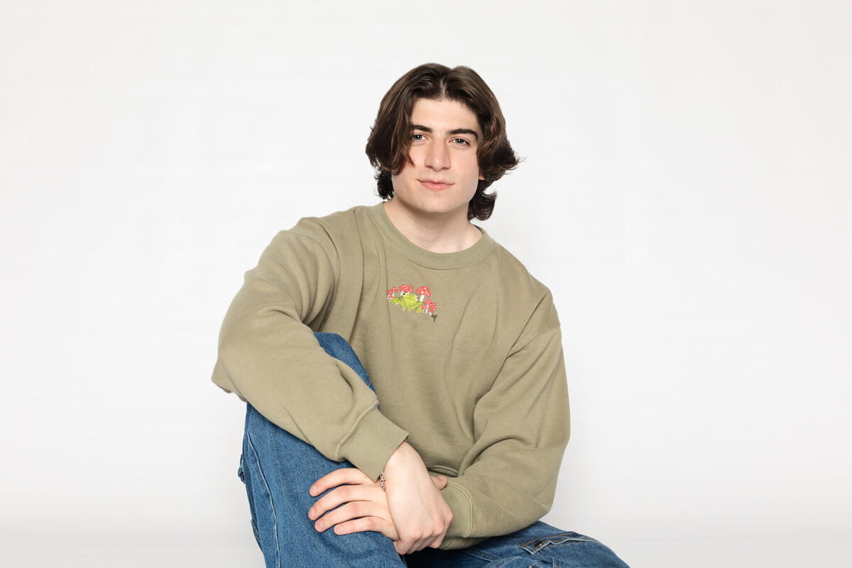 A young man with medium-length dark hair sits casually on the floor against a plain white background. He is wearing a light brown sweatshirt with a small graphic and blue jeans, looking relaxed.