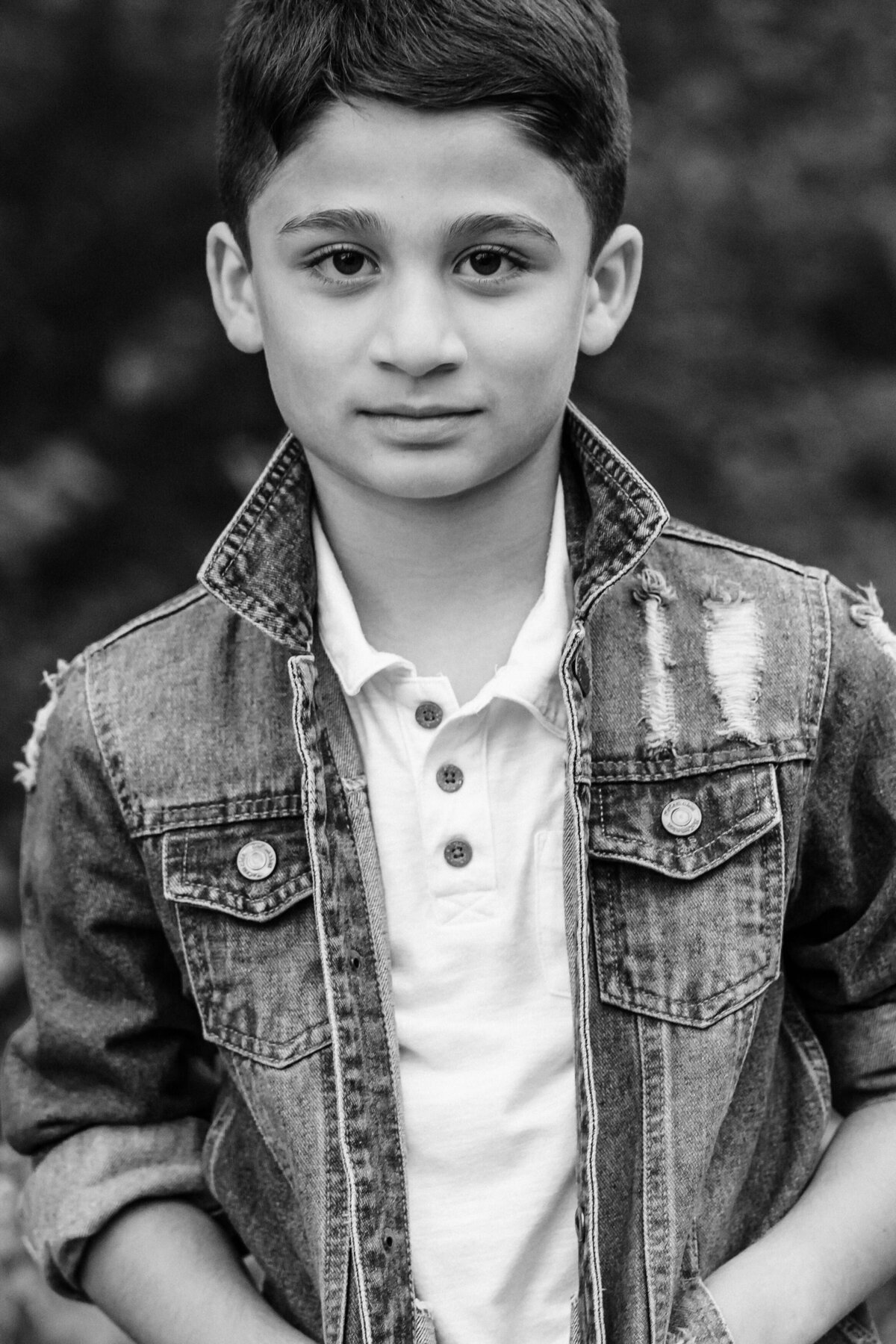 A close-up black and white portrait of a young boy looking directly into the camera, wearing a denim jacket over a white shirt, with a calm and composed expression.