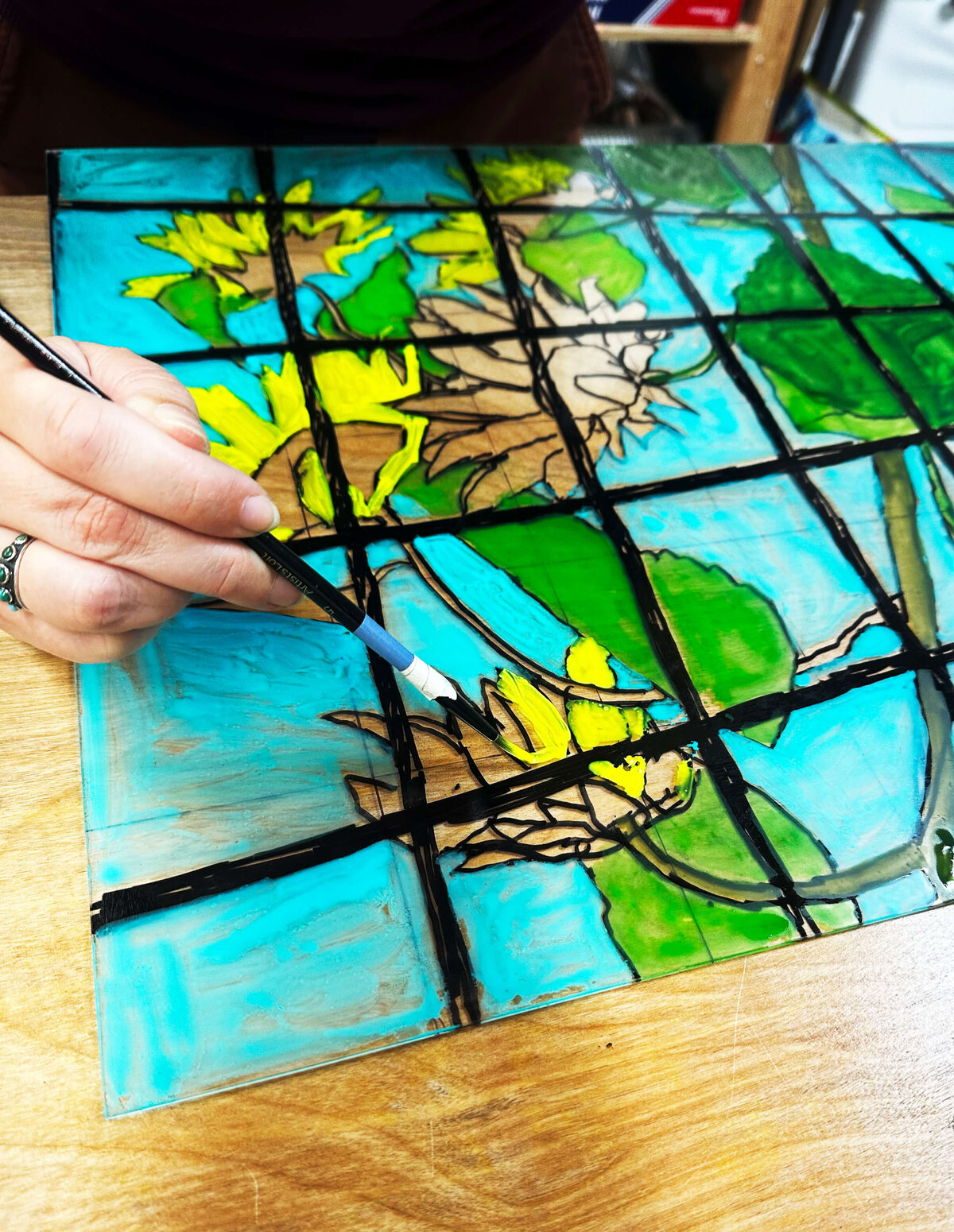 A close-up of a hand painting sunflowers in bold color on a gridded plexiglass plate, bringing the design to life before printing.