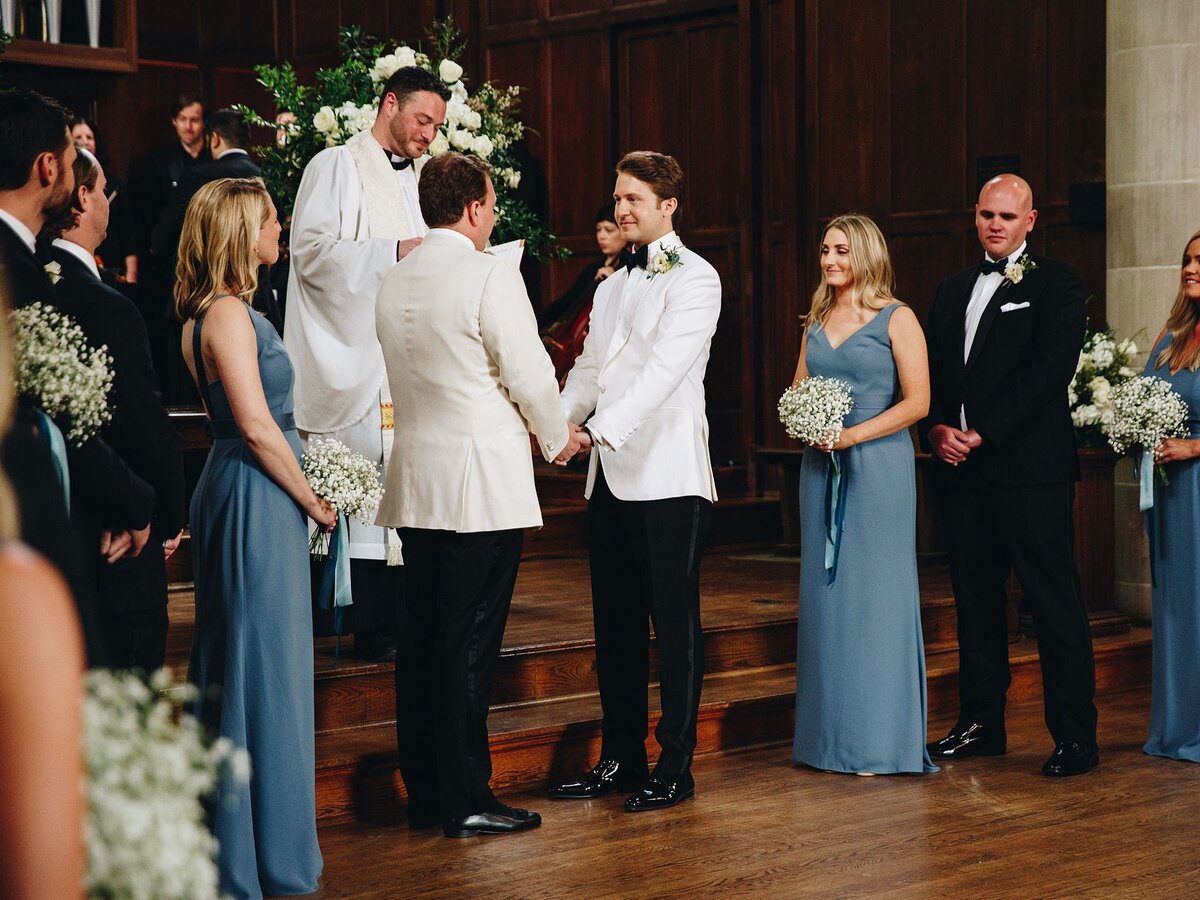 Two grooms wearing tuxedos with white jackets hold hands with their wedding party standing on either side wearing dusty blue dresses with large white floral arrangements in the background at Scarritt Bennett Center in Nashville