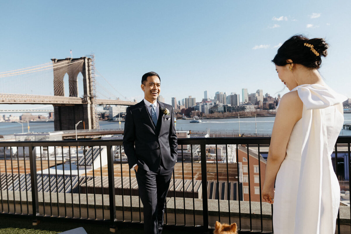 The bride and the groom smile at each other, as they stand on a terrace with a view of Brooklyn bridge and buildings.