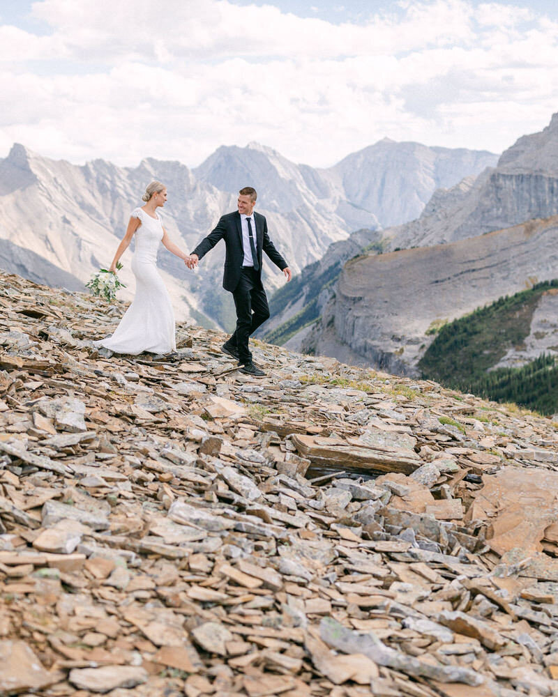 This lively photograph captures the bride and groom laughing and walking down a scree mountain slope during their helicopter adventure wedding in Banff National Park. The rugged landscape and their shared joy create a fun and adventurous moment on their unforgettable day.