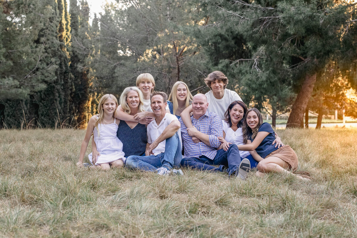 A large, extended family huddling together and sitting in puffy green grass at a park, surrounded by green trees and wearing shades of blue and white as they look at the camera. Captured at Jeffrey Open Space.