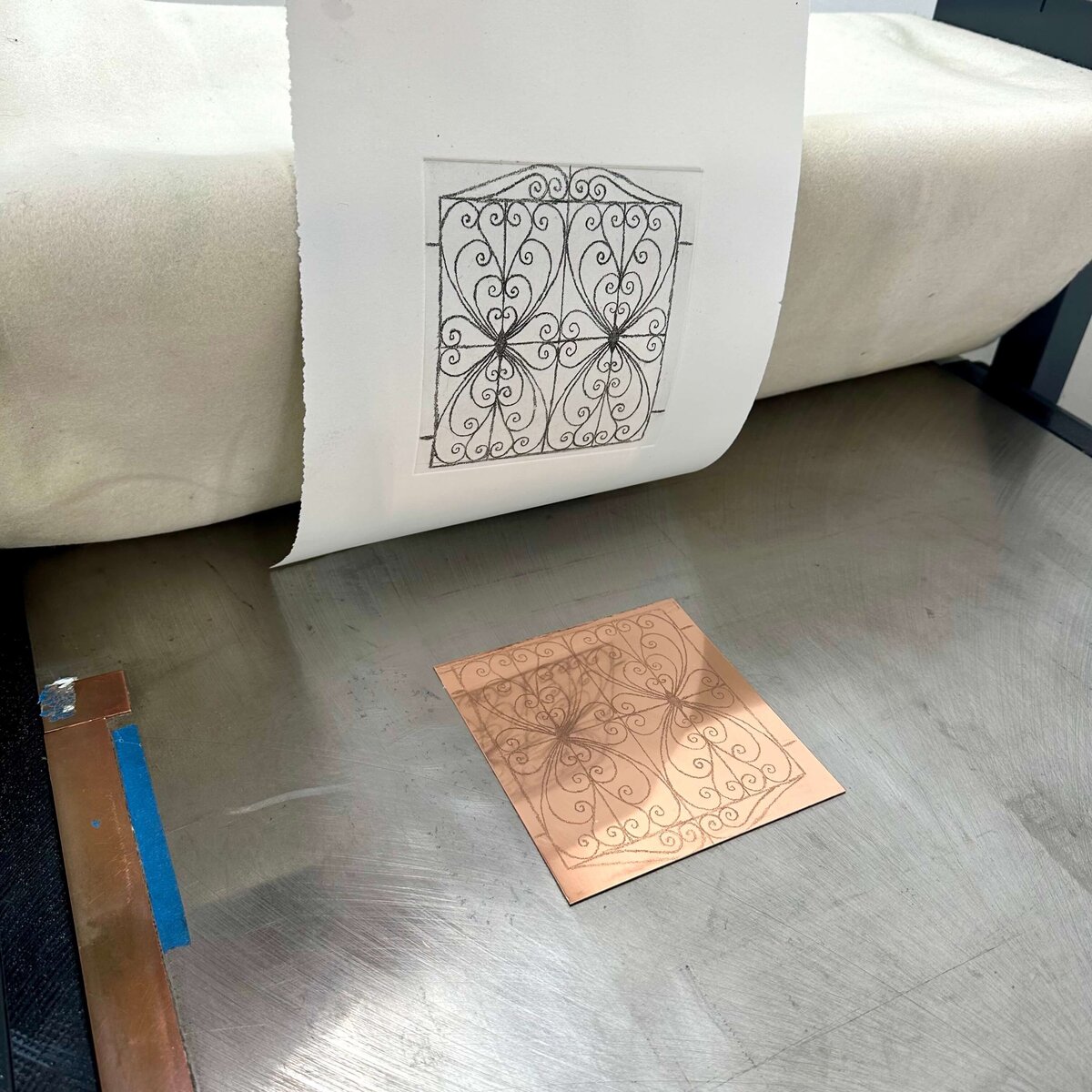 Copper plate and its matching print aligned under the press roller, capturing the printing process in motion.