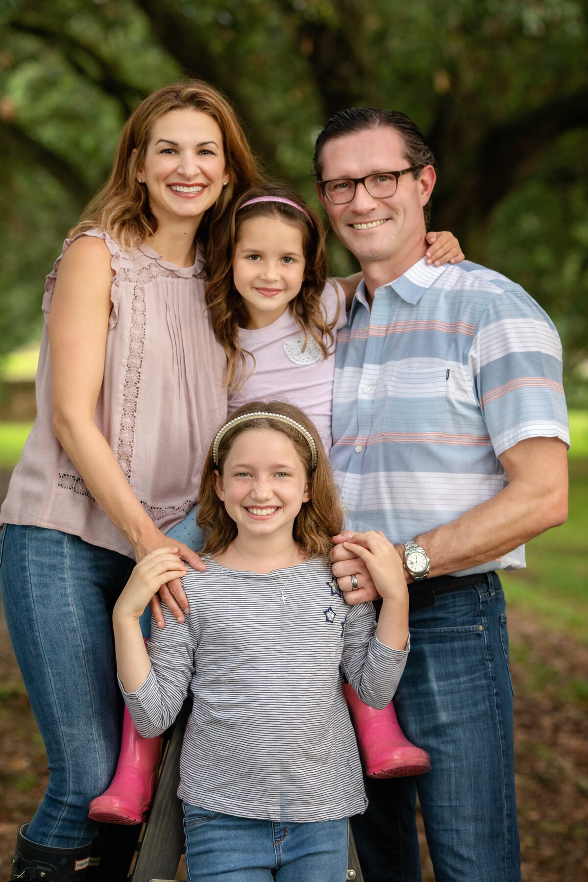 Family of 4 are posed together and looking at the camera.  There is a man, a woman, and 2 children.  One of the girls is wearing pink rainboots.  They are all smiling.