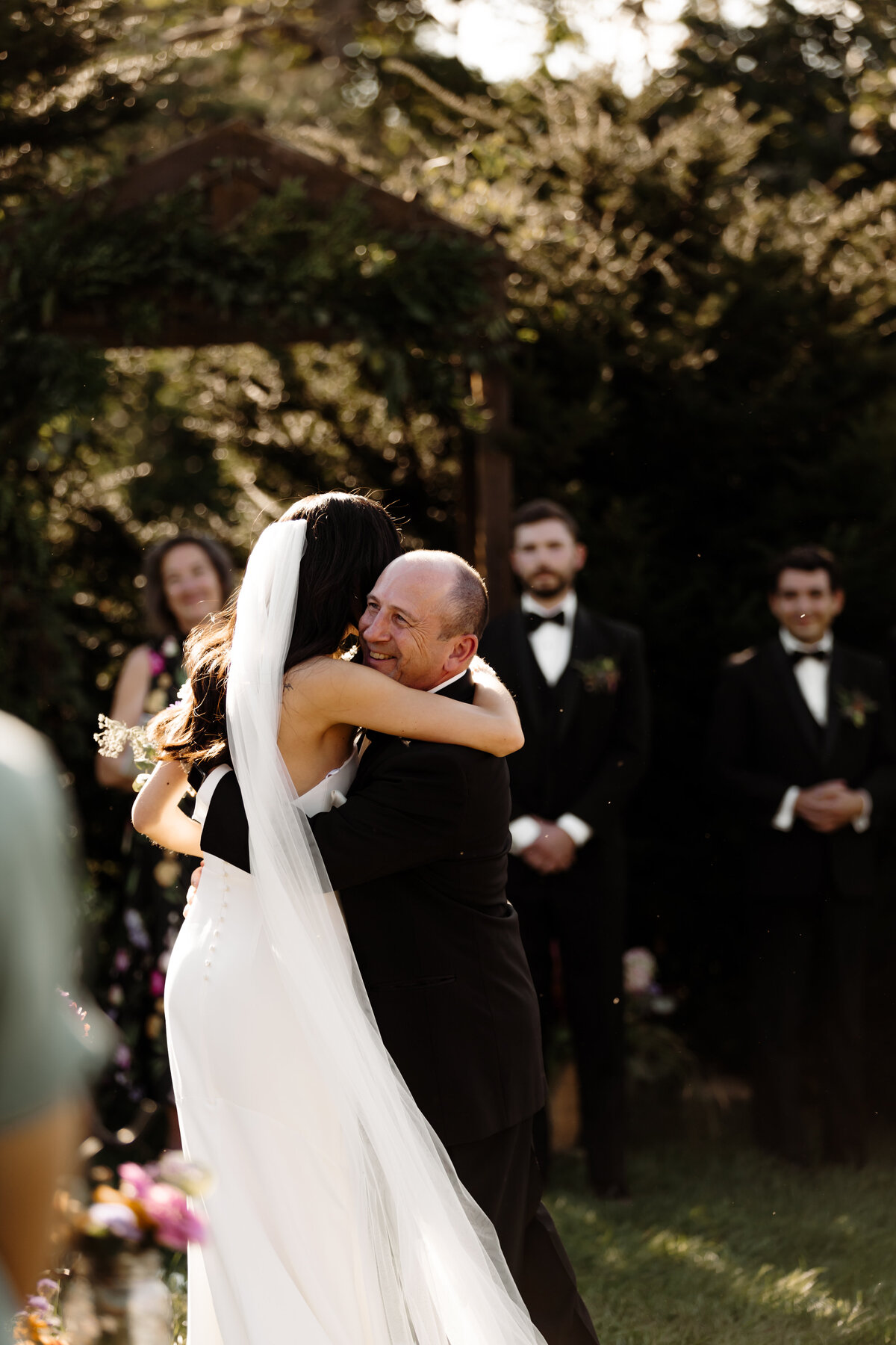 A bride hugs a man, both smiling warmly, during an outdoor wedding ceremony captured by a New England Wedding Photographer. She wears a long white dress and veil. In the background, guests in formal attire stand near trees and a wooden arch draped in foliage.