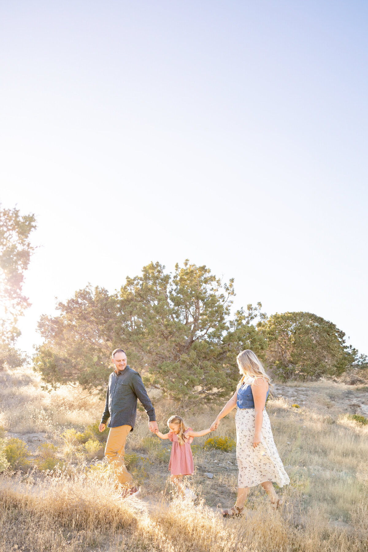 A family of three walks hand-in-hand along a sunlit path in Eagle Mountain, Utah. The low sun casts a golden glow, highlighting the desert landscape and sparse trees.