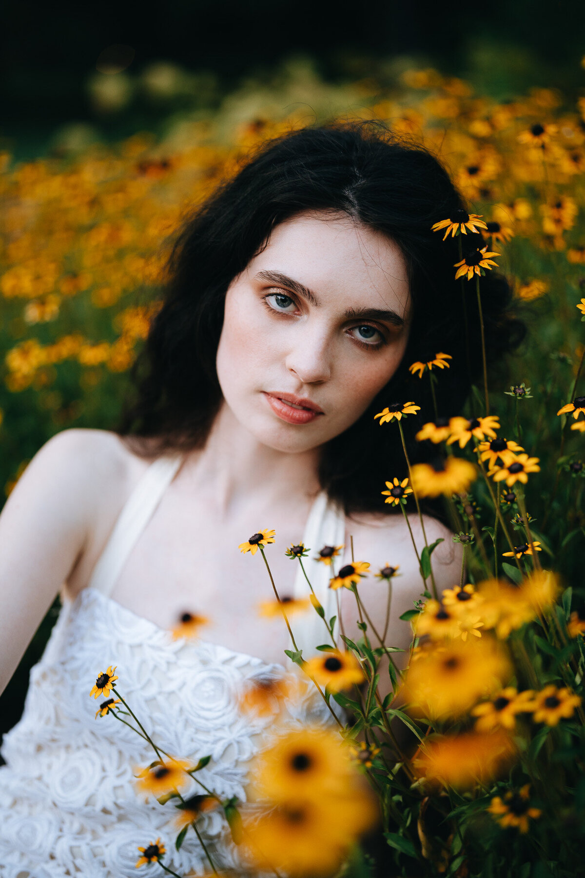 girl with dark hair wearing a white dress and sitting in a patch of yellow flowers
