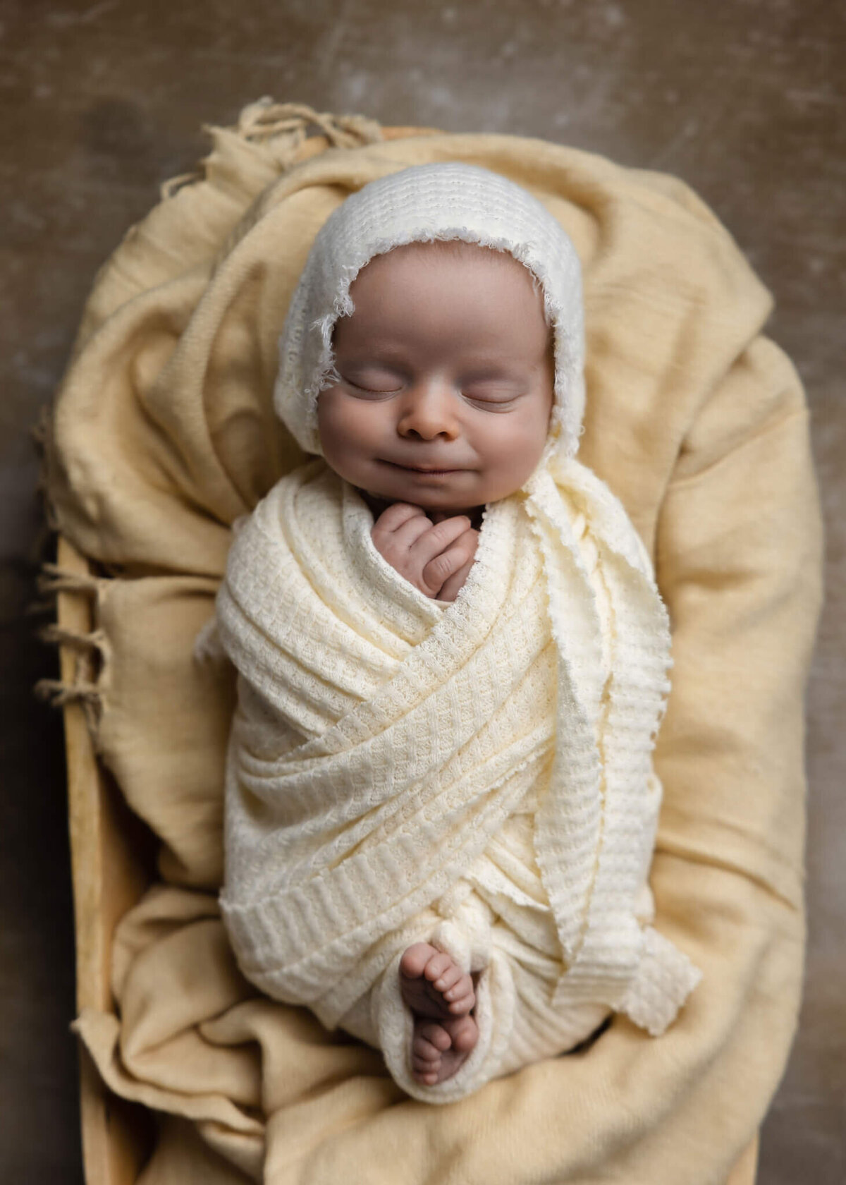 newborn baby wrapped in cream fabric and wearing a cream bonnet alseep in wooden bowl