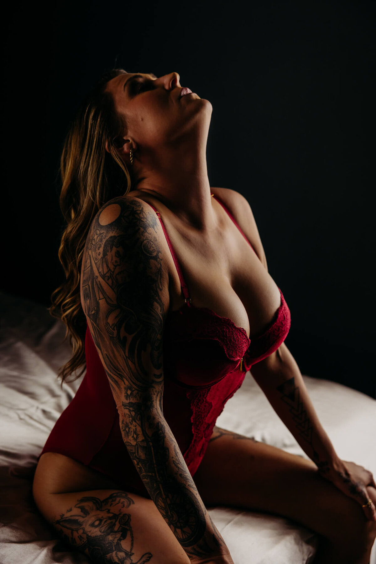 beautiful woman in a velvet red bodysuit sitting on the edge of a bed with her head back as she takes a breath in captured in a Oshkosh boudoir studio