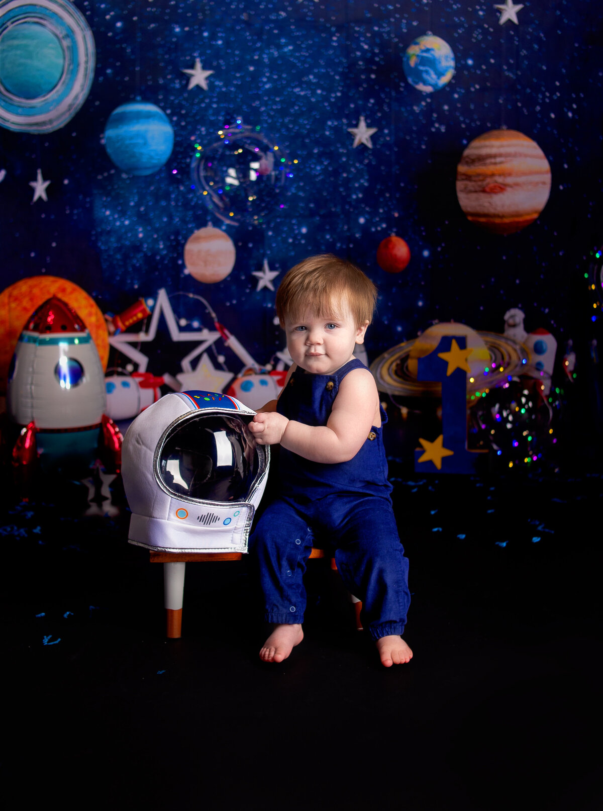 Little boy sitting on bench, holding space helmet during first birthday cake smash photoshoot in Franklin, TN photography studio with Franklin Tennessee family photographer