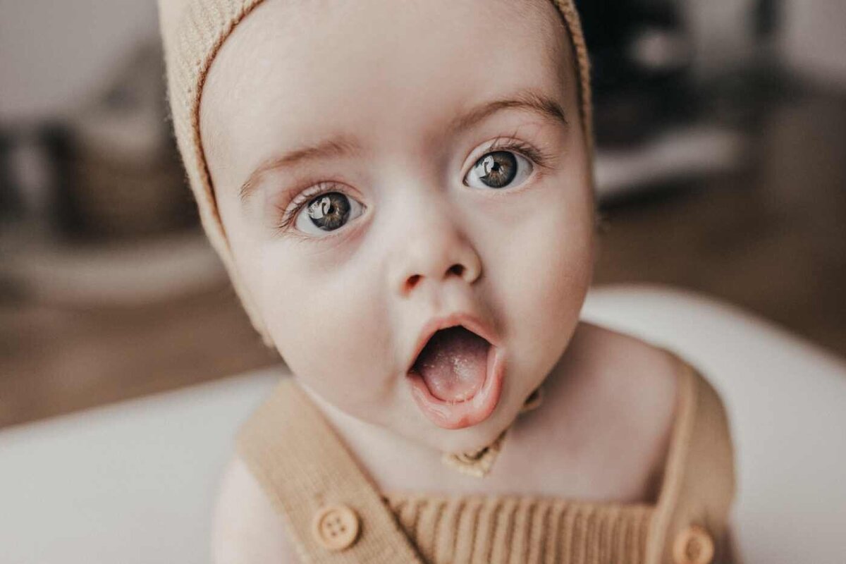 Ruua Hamdan Photography (Altrincham/Manchester, UK): A close-up of a baby with wide eyes and open mouth, wearing a beige outfit and bonnet.