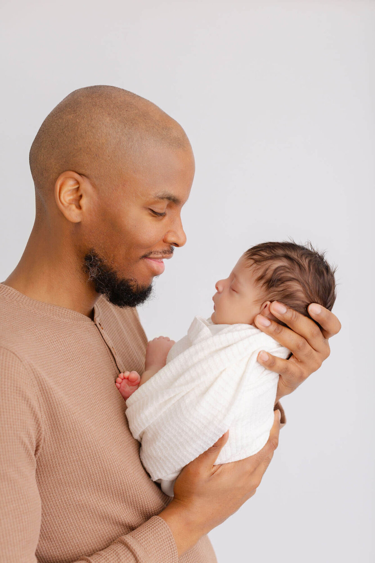 Dad in brown textured henley is holding newborn baby in front of him and smiling at him. This is a profile image of dad and baby. - PDX Newborn Photographer