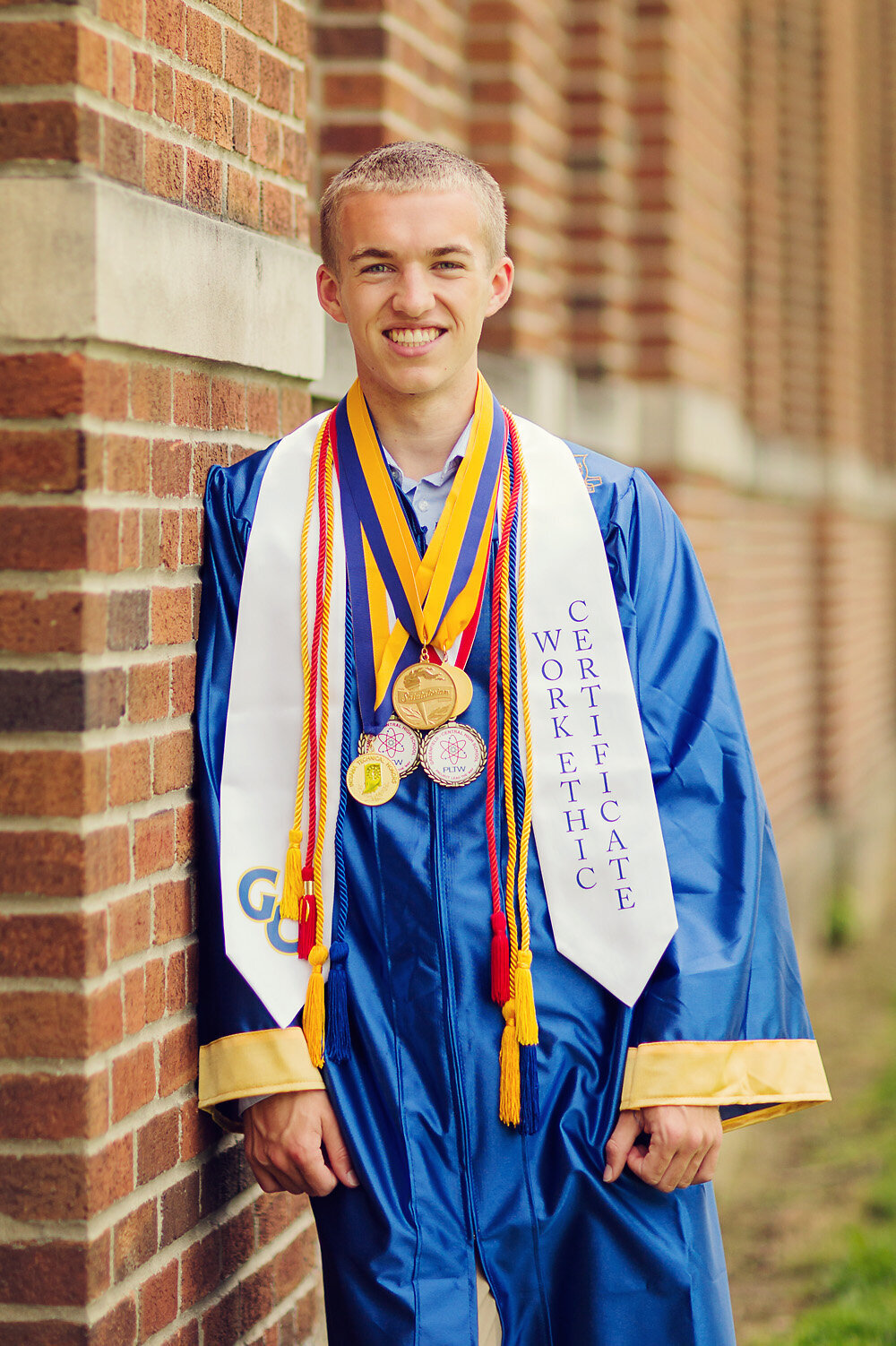 Connor in his high school gown and medals leaning against brick.