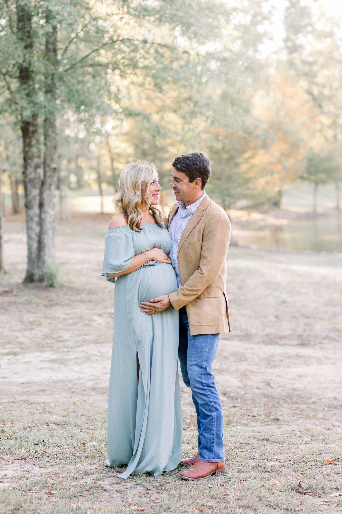 Maternity session with mom to be in blue dress standing with husband in a khaki jacket and blue jeans. Session location is on a pond bank. Picture taken by Brandon, MS Newborn photographer.