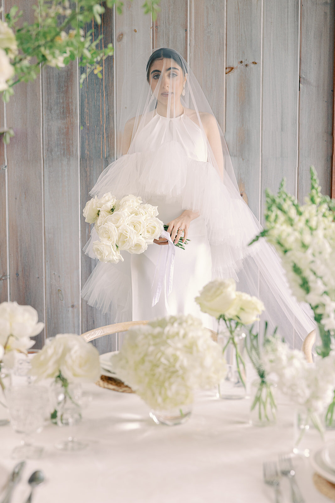 Bride with veil over her face holding a white bouquet in front of a table with white floral centerpieces