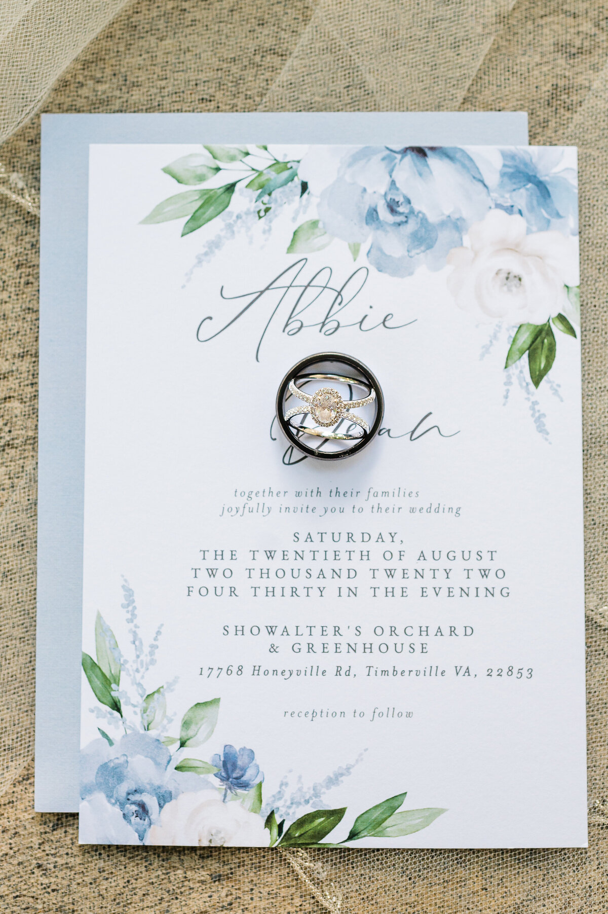 wedding invitation with light blue florals laying on a stone backdrop with the wedding rings of the bride and groom sitting on the top of invite