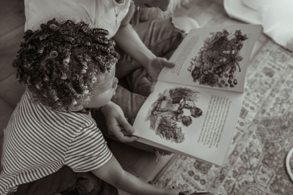 Overhead monochrome shot of a parent and child with curly hair reading a large picture book together on a patterned rug