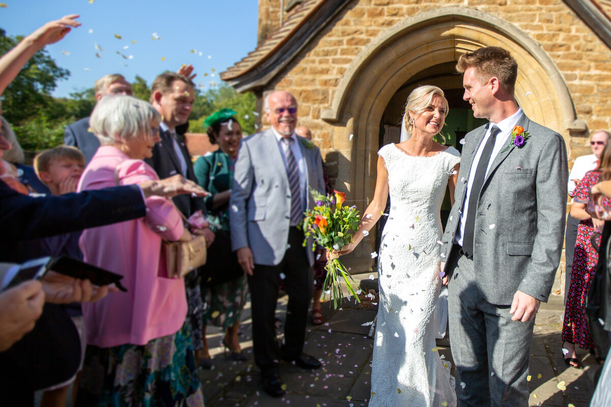 Happy couple leaving wedding chapel while guests throw petals