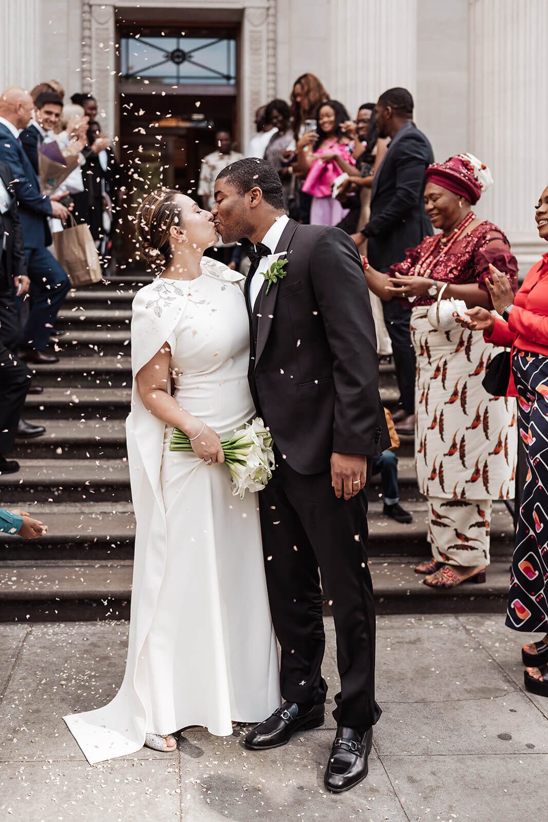 bride and groom kissing at the bottom of the steps outside marylebone town hall in london as their guests line the steps throwing confetti on them