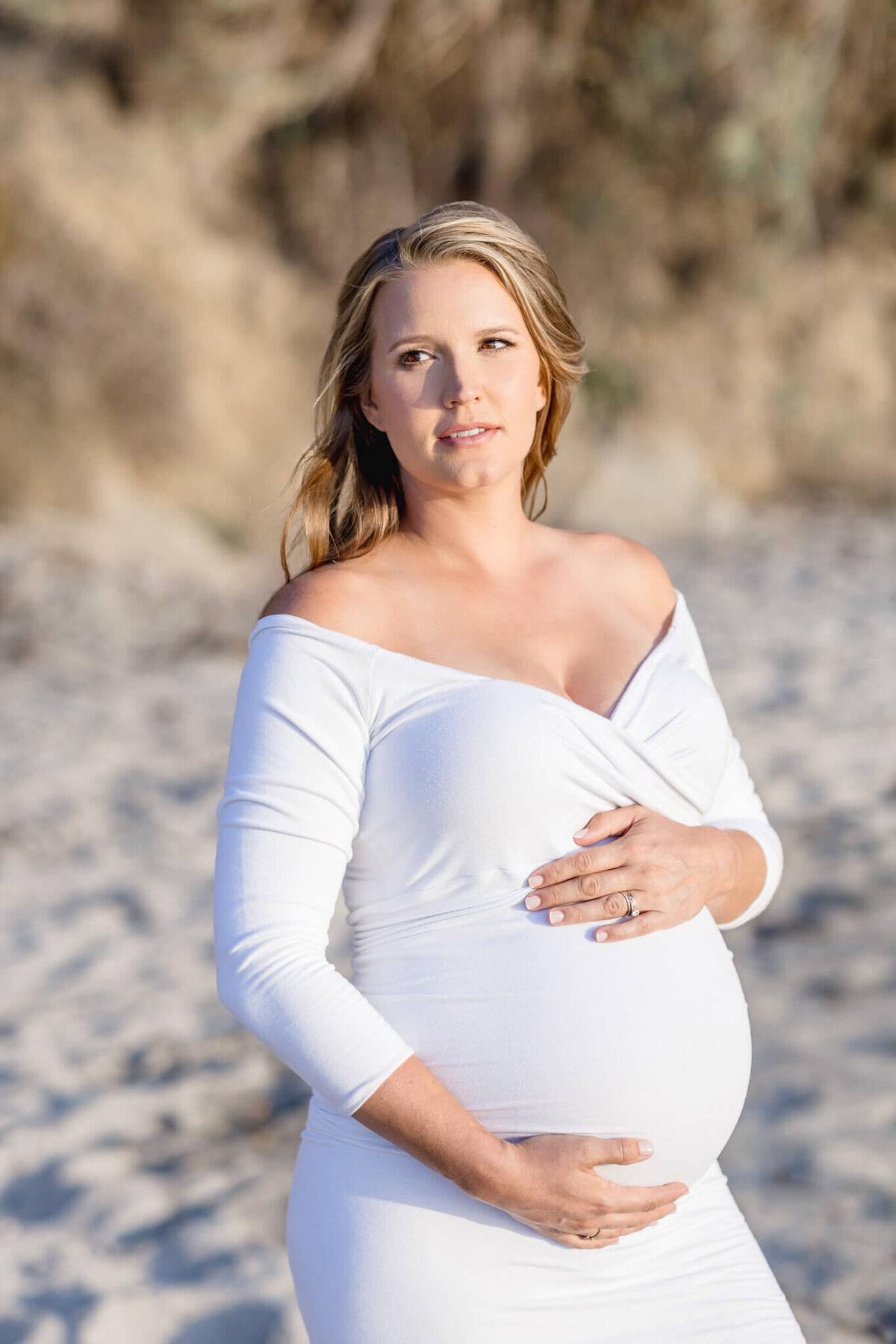 A pregnant woman in a white off-the-shoulder dress stands on a sandy beach, gently cradling her baby bump. She looks off into the distance with a serene expression. The backdrop is blurred with natural, earthy tones.