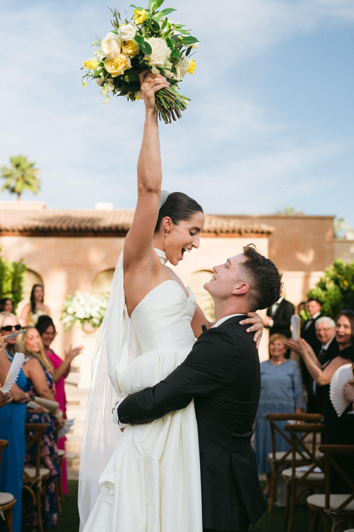 Groom lifts his bride in celebration as she raises her bouquet after the ceremony, captured by Arizona wedding photography team at Royal Palms.