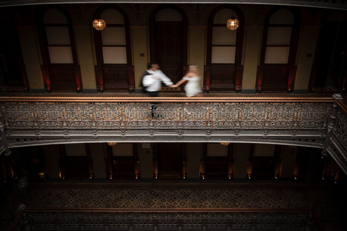 Blurred motion image of a couple holding hands while walking across the ornate upper-floor balcony of the Beekman Hotel; the photo highlights the cast iron railings, rich wood paneling, and warm lighting—an artistic example of Beekman Hotel engagement photos that captures both movement and architectural detail.