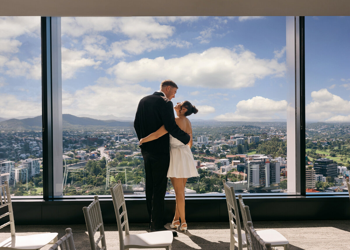Couple embracing in front of floor-to-ceiling windows at the Brisbane Registry Office, with panoramic views of the city skyline.
